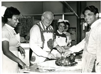 A photograph of two doctors in aprons carving a turkey on a hospital ward. There are also three nurses in uniform in the photograph, either side of the doctor in the middle.