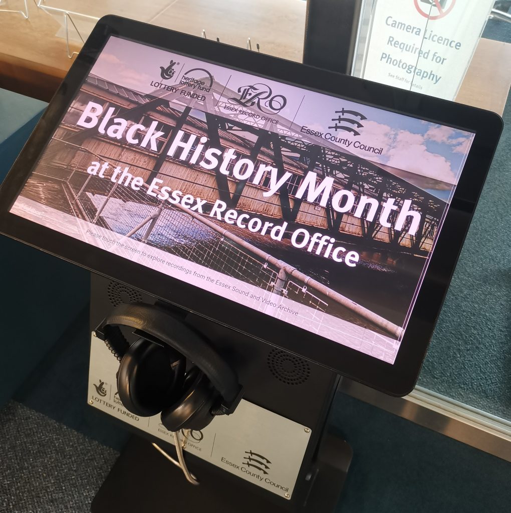 Photograph of a listening post in the Searchroom at the record office. The main screen has an image of an art installation in a walkway at the Port of Tilbury, with text overlaid that reads: "Black History Month at the Essex Record Office". Below the main screen is a pair of headphones.