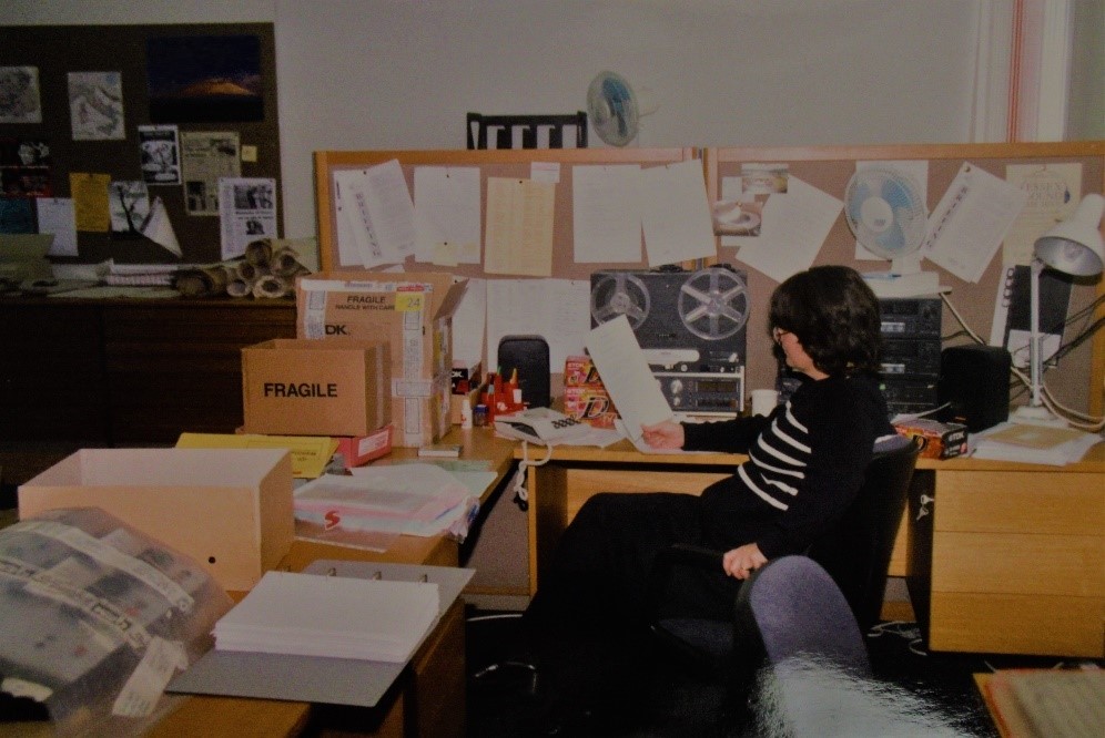 Photograph of an office, with a corner desk and pinboard in the background. A woman, Sue Cubbin, is sitting side on at the desk, looking at a piece of paper in front of a reel-to-reel tape machine.