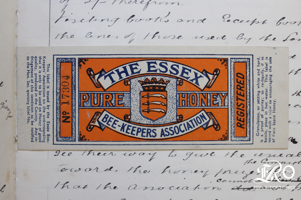 A thin cardboard label designed to wrap around a glass honey jar. Heraldic design with The Essex Beekeepers' Association" and "Pure Honey" written in banners around the Essex county coat of arms