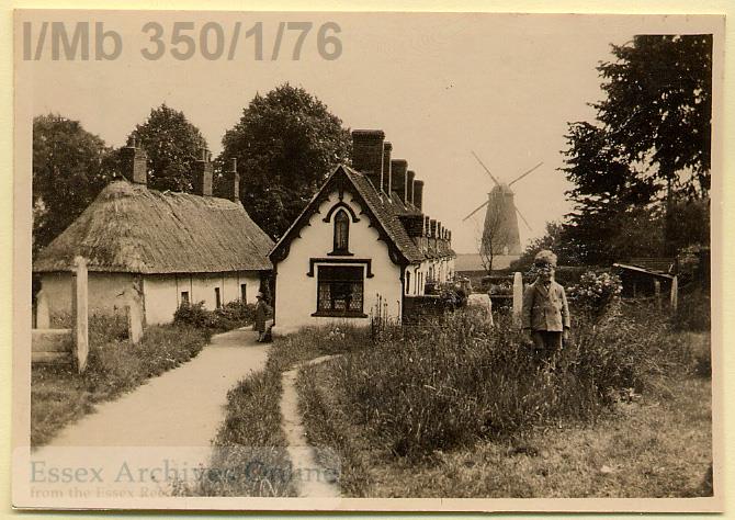 Black and white photograph of Thaxted chantry with a thatched roof on the left and almshouses on the right with John Webb's windmill, also known as Lowe's Mill, in the background. A young boy is standing in the midground on the right wearing shorts and a jacket