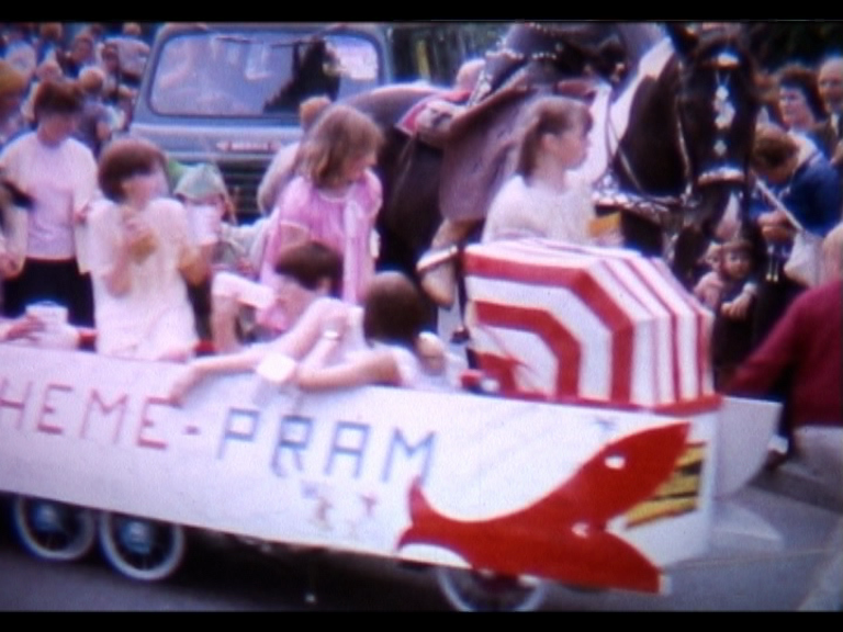 Slightly fuzzy still from a cine-film, showing several prams linked together to create a carnival float. The pram at the front has a red and white striped hood, and there is a sign across the prams reading 'Theme-Pram'. There are children in the prams and behind in the crowds, along with a horse and another vehicle.