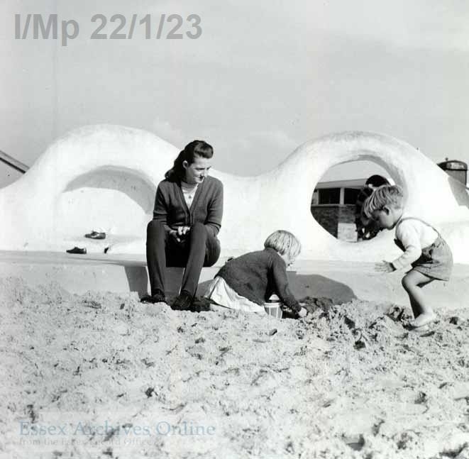 Black and white photograph of a woman with two young children playing in a sand pit