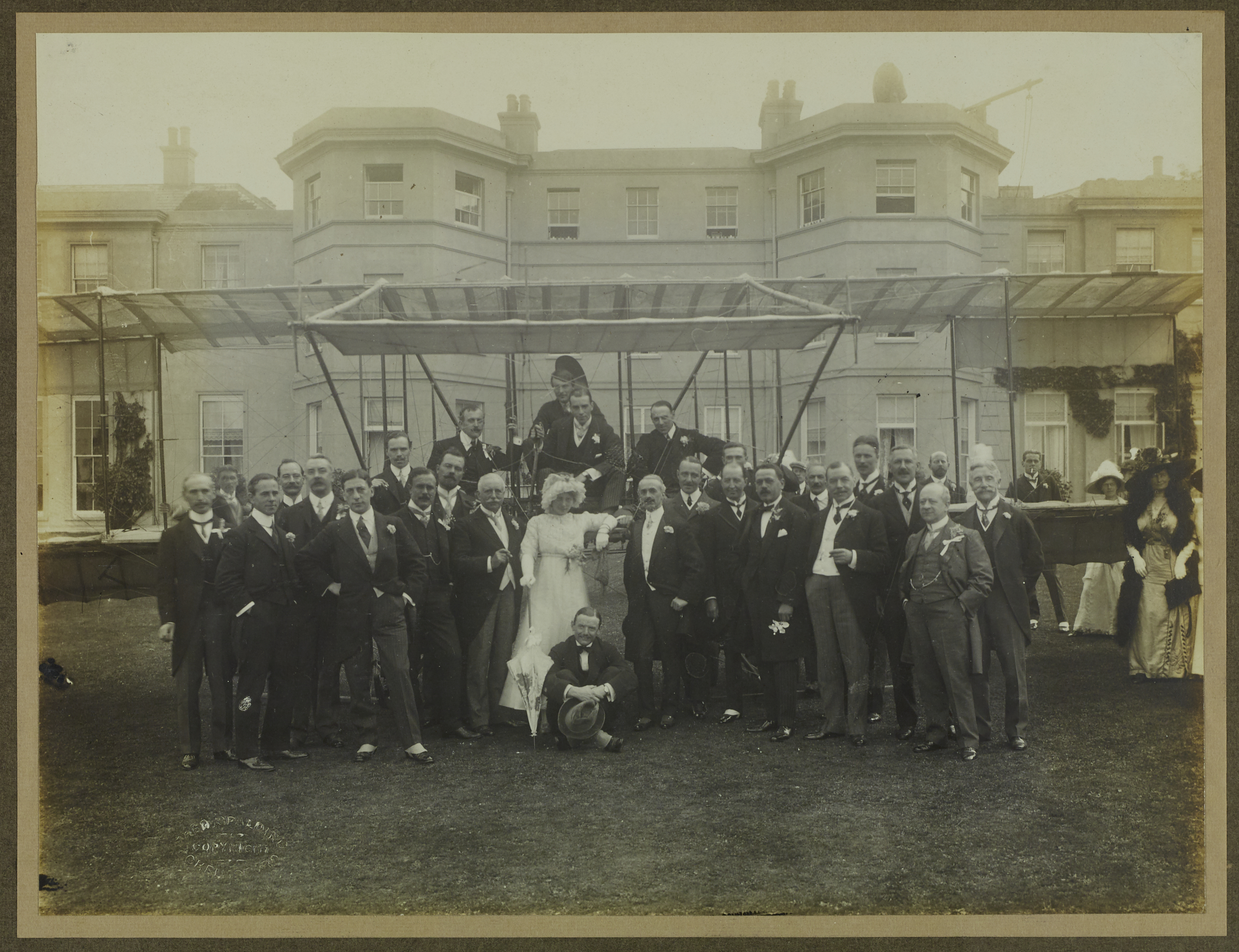 Black and white photograph of wedding party (mainly men with some women nearly out of shot) posing with a biplane in front of Hylands House. Groom and several men on the biplane wing. Bride standing in front of the biplane in white wedding dress