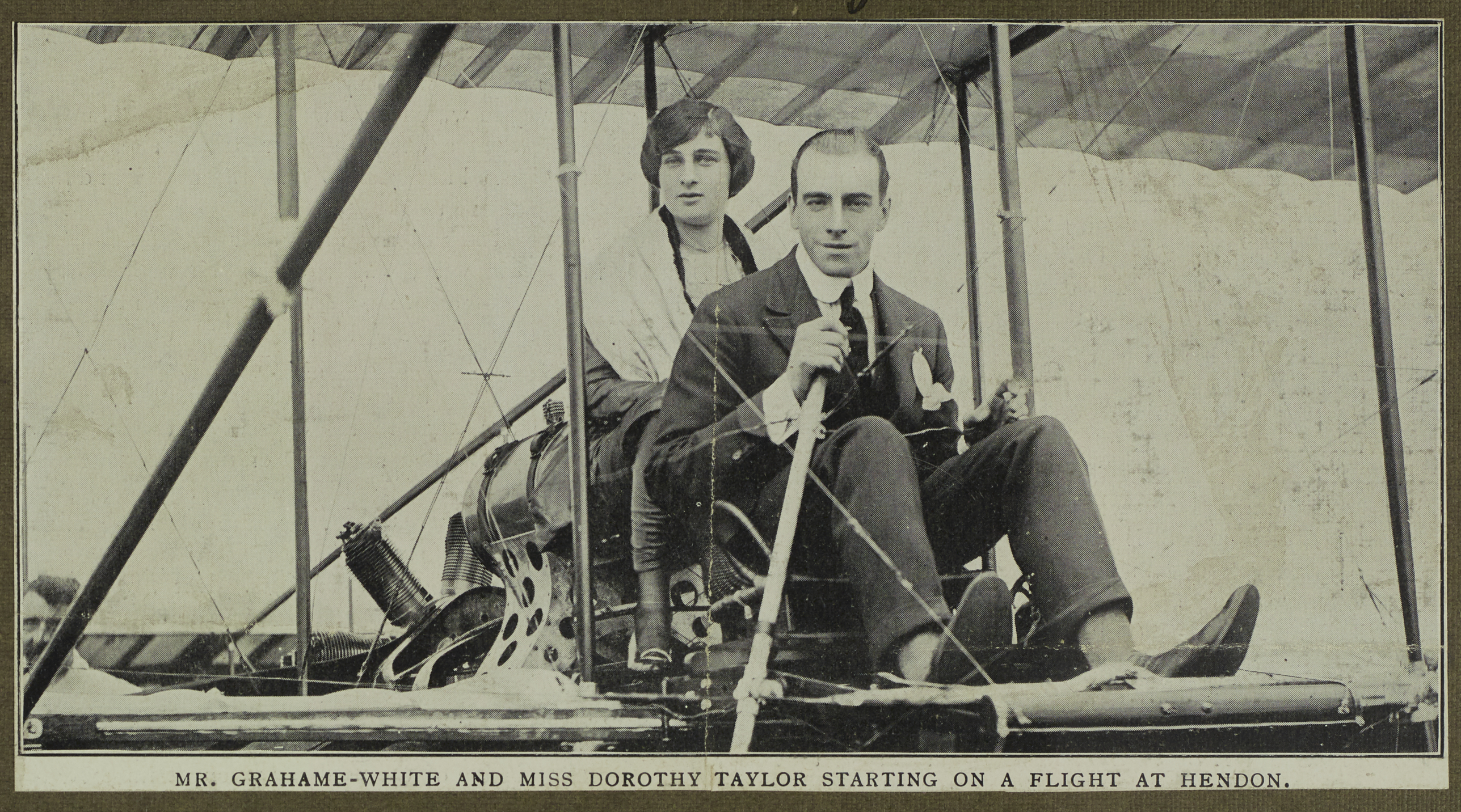 Black and white photograph of a man (Mr Grahame-White at the front) and woman, Miss Dorothy Taylor sitting in a biplane. The man is wearing a suit and tie and the woman is wearing a dress or skirt and shawl.