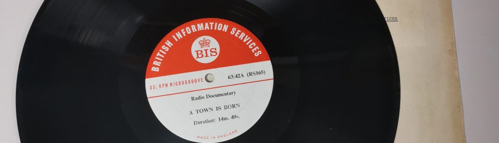 Photograph from above of vinyl disc, with red 'British Information Services' label. Laid on top of yellowed disc sleeve on white surface.