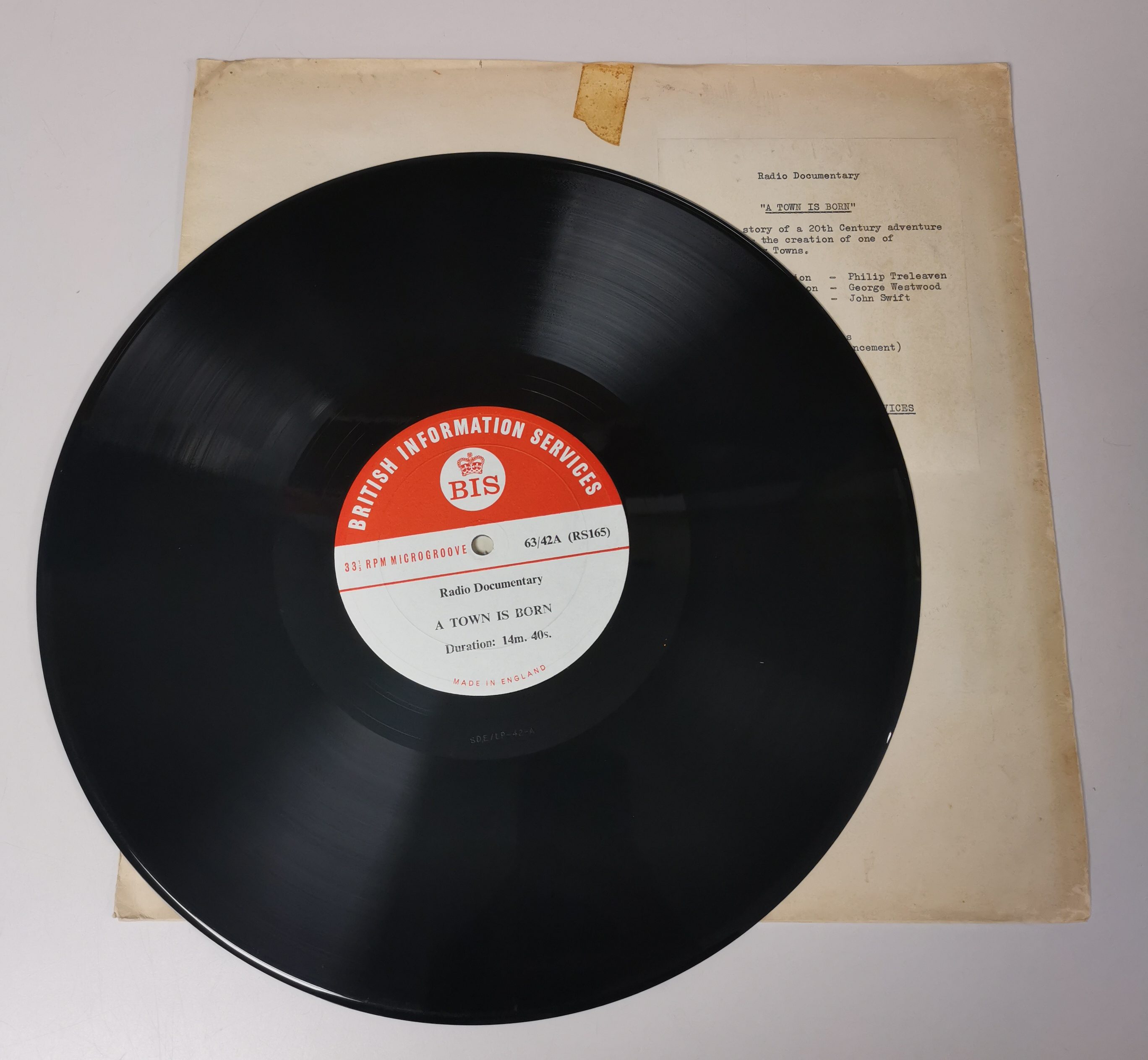 Photograph from above of vinyl disc, with red 'British Information Services' label. Laid on top of yellowed disc sleeve on white surface.