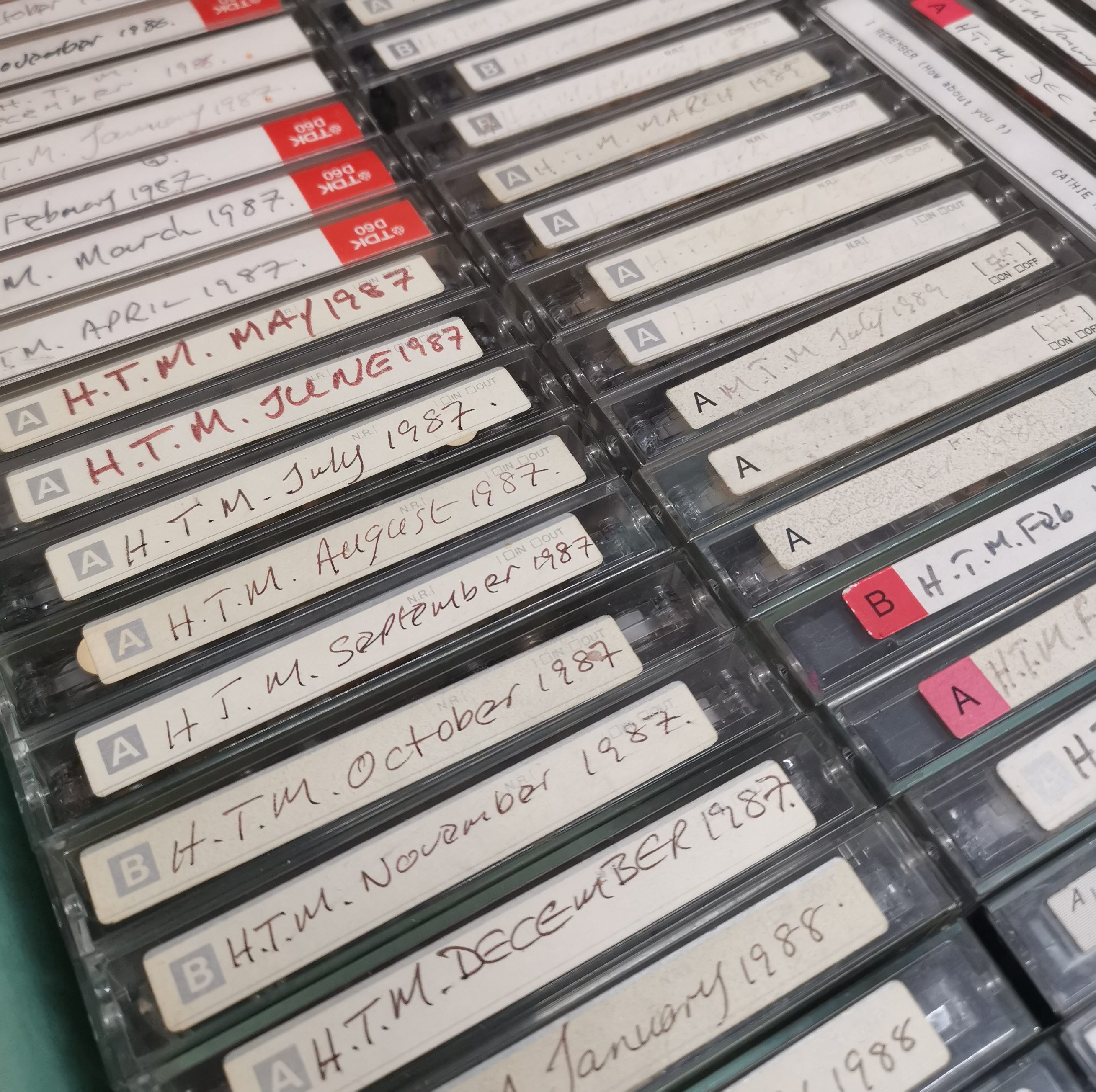 Three rows of cassettes in a green box. Handwritten labels on the cassettes read 'HTM' and the month and year they were created.