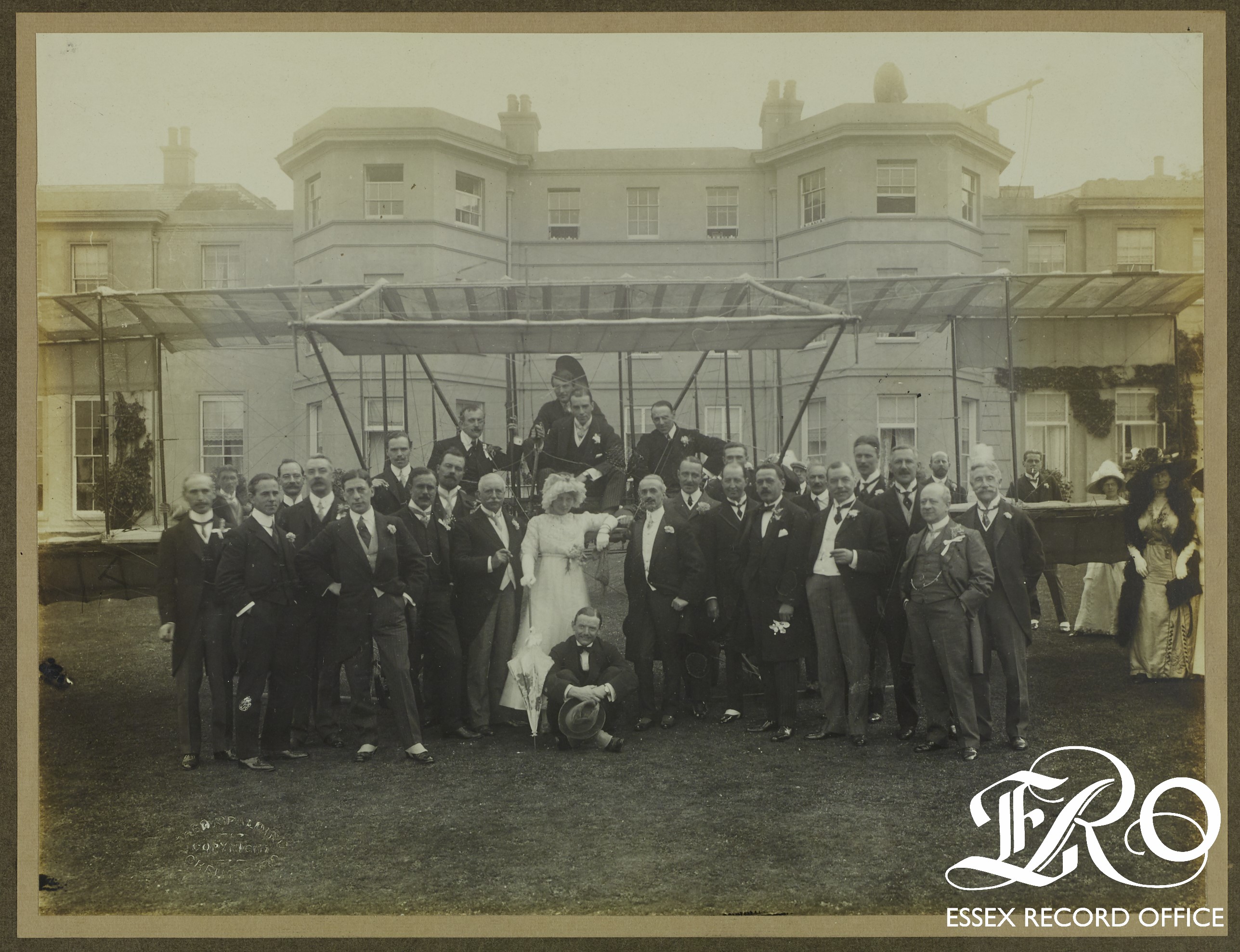 Black and white photograph of wedding party (mainly men with some women nearly out of shot) posing with a biplane in front of Hylands House. Groom and several men on the biplane wing. Bride standing in front of the biplane in white wedding dress